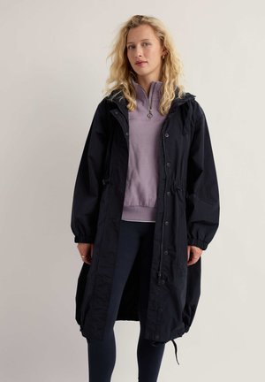 Blonde vrouw draagt een lange zwarte regenjas over een lichtpaarse zip-up top en donkere legging, staand tegen een effen beige achtergrond.