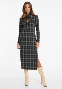 Langärmliges, schwarz-grau kariertes Kleid mit hohem Kragen, versehen mit einem Adlerdesign auf der Brust und einem seitlichen Schlitz; kombiniert mit cremefarbenen kniehohen Stiefeln.