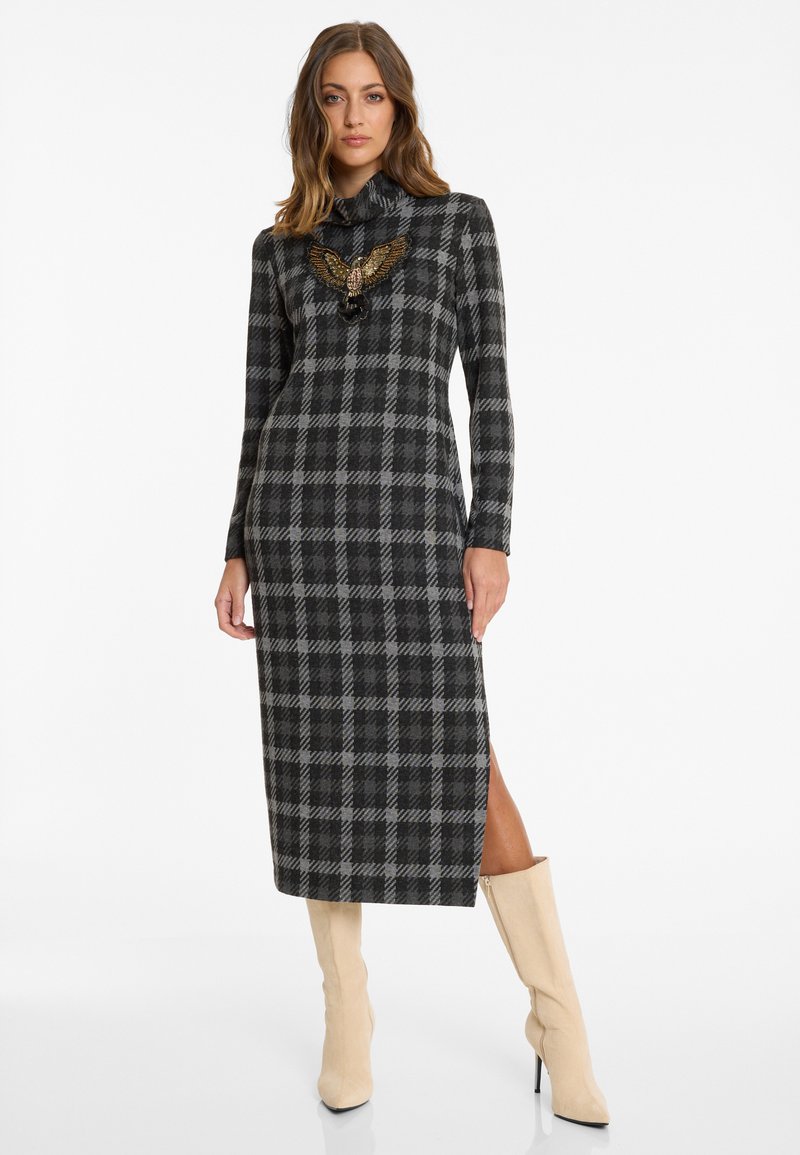 Langärmliges, schwarz-grau kariertes Kleid mit hohem Kragen, versehen mit einem Adlerdesign auf der Brust und einem seitlichen Schlitz; kombiniert mit cremefarbenen kniehohen Stiefeln.