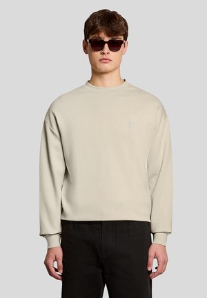 Young man wearing beige sweatshirt with small embroidered logo, black pants, and dark sunglasses, standing against plain background.