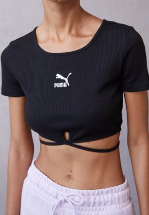 Personne portant un t-shirt court noir Puma avec un petit œillet et un détail à nouer, associé à un pantalon rose clair à cordon de serrage.