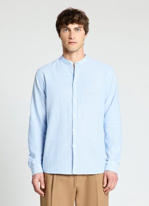 Homme portant une chemise boutonnée à rayures bleu clair avec col mandarin et un pantalon beige, debout sur un fond clair uni.