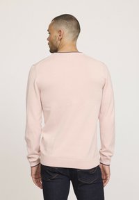 Pull en tricot rose clair à manches longues, col rond et poignets côtelés. Porté par-dessus un jean en denim foncé. Design minimaliste sans motifs visibles.