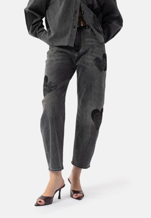 Grå denimjeans med svarte teksturerte hjerteapplikasjoner på knærne, kombinert med svarte åpne tåhæler og en løs grå skjorte med knapper.