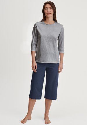 Vrouw staat blootsvoets, draagt een gestreept shirt met lange mouwen en wijde cropped broek in marineblauw op een effen lichte achtergrond.
