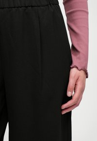 Pantalons noirs avec une texture lisse, une taille élastique et deux plis verticaux à l'avant. Un haut rose à rayures à manches longues dépasse sur le côté.