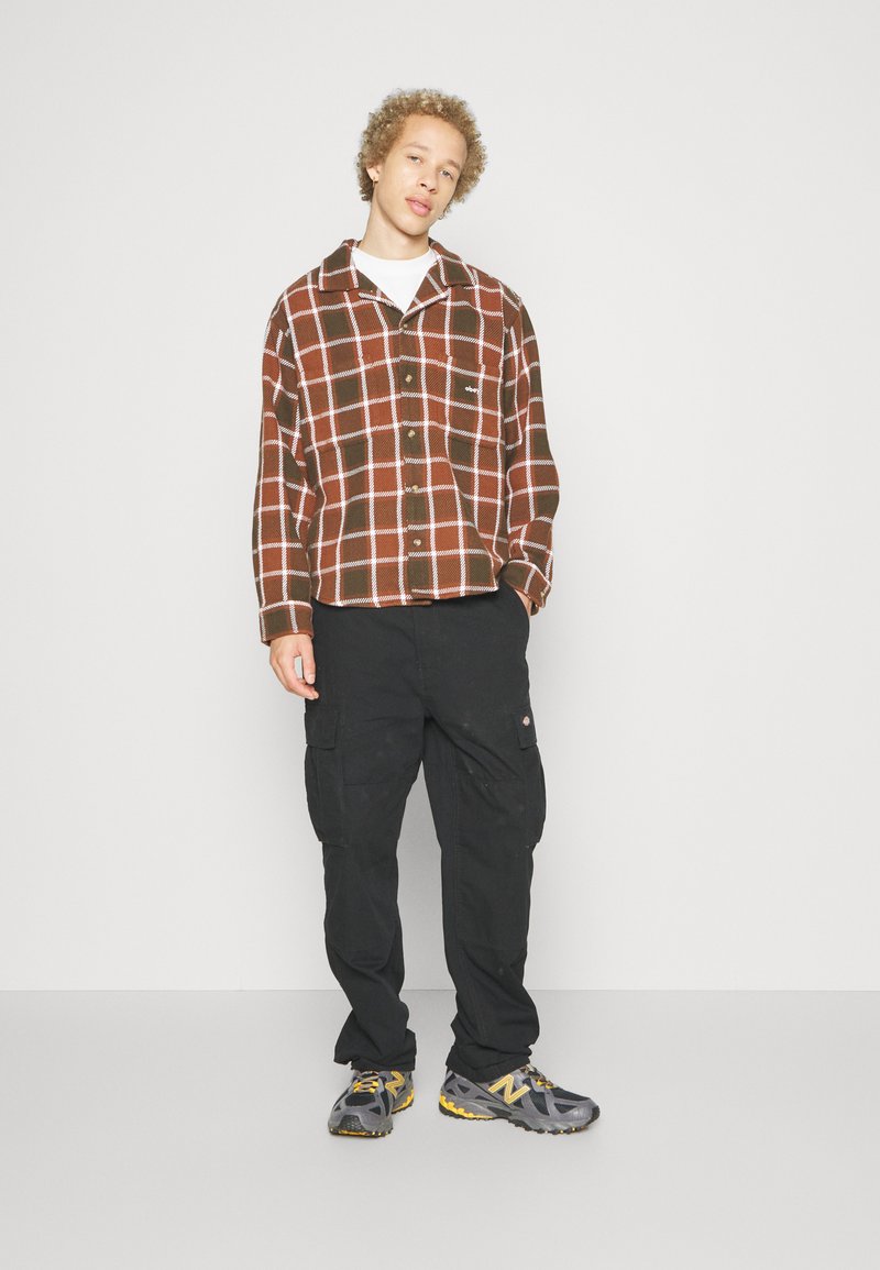 Camicia a quadri marrone e bianca in flanella, pantaloni cargo neri e sneakers grigie con accenti gialli. Outfit casual con una vestibilità morbida.