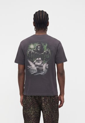 Άτομο με πλεξούδες που φοράει σκούρο T-shirt με γραφικό ενός Θανάτου και ενός πλοίου, σε συνδυασμό με παντελόνι με λεοπάρ σχέδιο, γυρισμένο πλάτη.