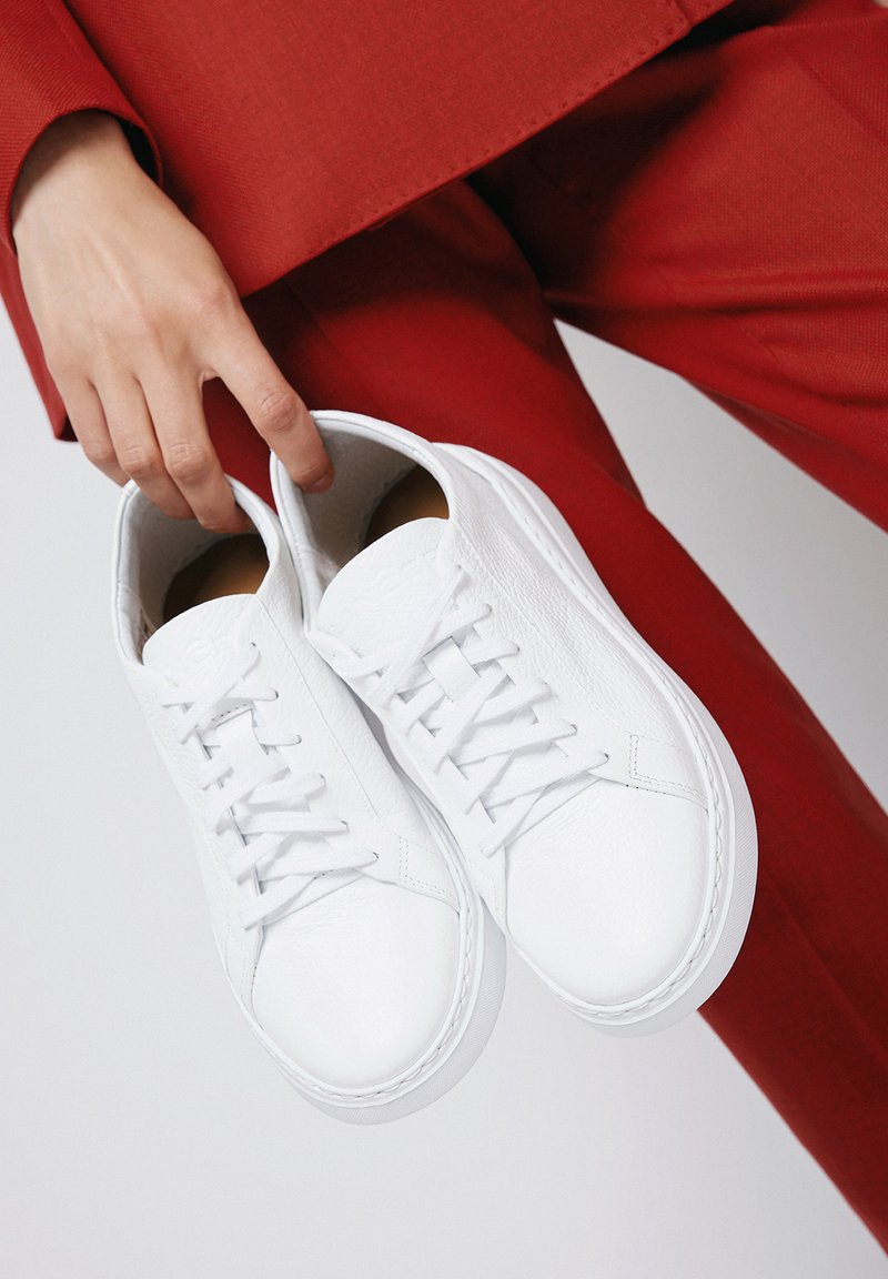Białe skórzane trampki z okrągłym noskiem, teksturowaną powierzchnią i płaskimi sznurowadłami. Podeszwa jest biała z lekkim podniesieniem. Trzymane na czerwonym tle.