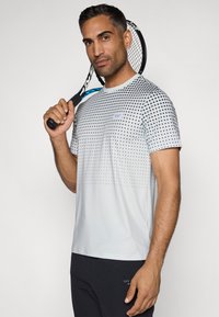 Lichtgrijs sportshirt met stippenpatroon, ronde halslijn, korte mouwen en een label op de borst. Zwarte broek en tennisracket zijn inbegrepen.