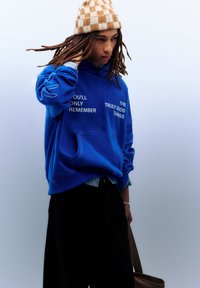 Blauwe pullover hoodie met witte tekst op de voorkant. Voorzien van een voorzak, geribbelde manchetten en een losse pasvorm. Draagt zwart broek en een gebreide muts.