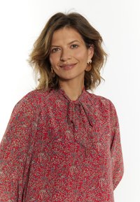 Bluză florală roșie cu gât legat, având modele intricate de frunze și flori în nuanțe de albastru și crem. Material ușor și croială lejeră.