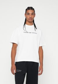 T-shirt branca de algodão com mangas curtas, apresentando o texto impresso em preto "TEMPERATURA DO SISTEMA. CRÍTICA" na parte frontal; modelo a usar calças pretas.