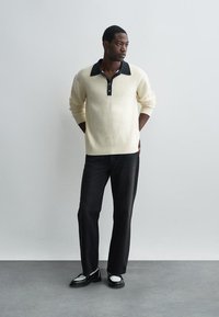 Cremfarbener Strickpullover mit schwarzem Kragen und Knöpfen, kombiniert mit dunkelblauen Jeans und schwarzen Schuhen mit weißen Akzenten. Einfaches, strukturiertes Design.