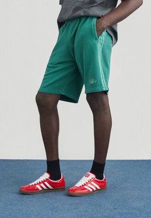 Pantaloni scurți verzi din bumbac cu buzunare laterale, cu croială lejeră și talie cu elastic striat. Asortați cu adidași roșii și șosete negre până la glezne.