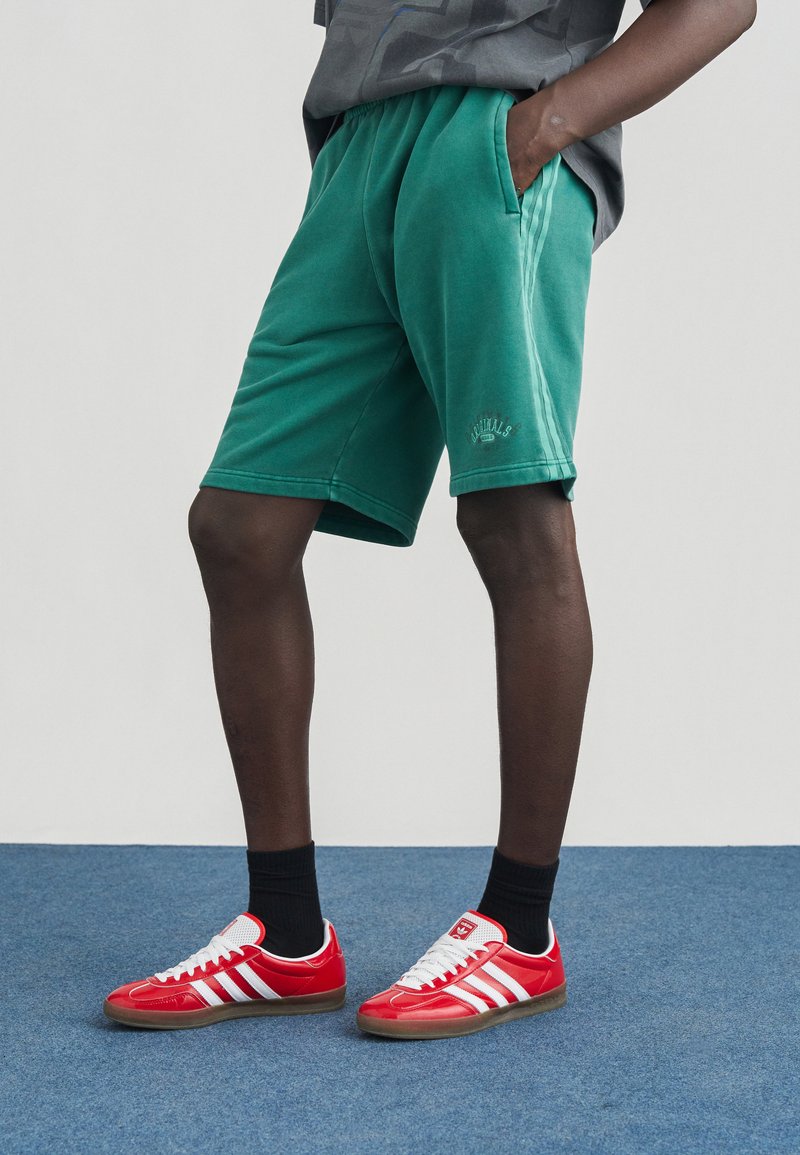 Πράσινα βαμβακερά σόρτς με πλαϊνές τσέπες, άνετη εφαρμογή και ribbed ελαστική μέση. Συνδυάζονται με κόκκινα αθλητικά παπούτσια και μαύρες κάλτσες μέχρι τον αστράγαλο.