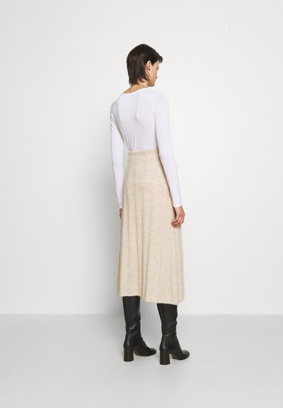 Saia midi de malha creme com textura canelada e cintura alta. Combinada com botas pretas de salto, a roupa apresenta um top branco de mangas longas.