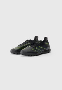 Une paire de chaussures de soccer noires avec des accents vert néon et des semelles texturées, conçues pour jouer sur gazon ou herbe artificielle.
