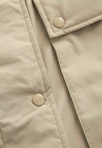 Gros plan sur le tissu d'une veste matelassée beige avec des coutures renforcées et deux boutons-pression ronds sur des panneaux superposés.