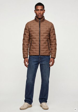 Mann trägt eine braune gesteppte Jacke, blaue Jeans und beige Sneaker und steht vor einem schlichten hellen Hintergrund.