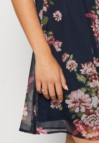 Marinblommig klänning med genomskinlig överdel, prydd med rosa och vita blommor. Hand synlig, bär en guldring med en liten sten på fingret.