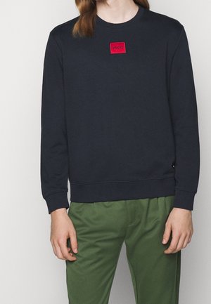 Osoba v černé mikině s dlouhým rukávem s červenou nášivkou "HUGO" a olivově zelenými kalhotami, stojící před jednobarevným pozadím.