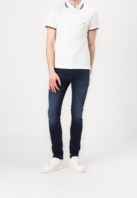 Vit polo med marinsvart och röd randig krage, i kombination med mörkblå skinny jeans och vita sneakers, står mot en neutral bakgrund.