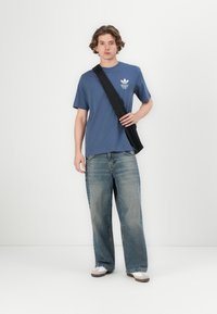 Blå bomulds t-shirt med korte ærmer og hvidt logo, parret med løstsiddende lyseblå jeans. Sort sling-taske båret over kroppen.