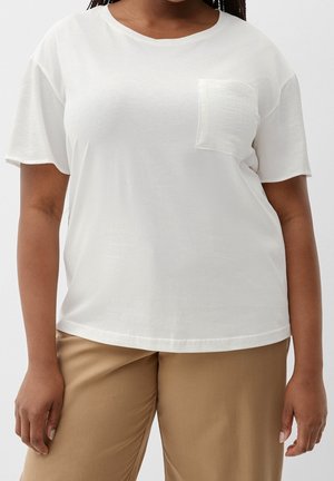 Vrouw die een wit kort mouw shirt met een borstzak draagt en beige broek, afgebeeld van nek tot halverwege de dij tegen een effen achtergrond.
