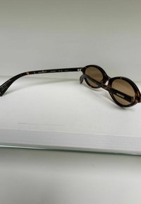 Lunettes de soleil rondes à monture écaille de tortue avec verres bruns, posées sur une surface blanche, une branche étendue et marquée Komono.