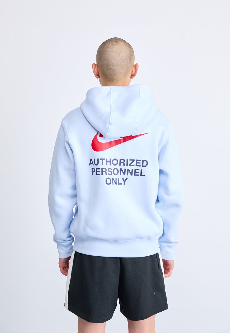 Világoskék kapucnis pulóver piros Nike logóval és a hátán "CSAK ENGEDÉLYEZETT SZEMÉLYEK" felirattal. Fekete rövidnadrággal párosítva.