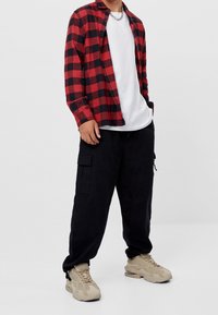 Rood-zwart geruit overhemd van flanel, gedragen over een wit T-shirt, gecombineerd met zwarte cargobroek en beige sneakers. Accessoire van een zilveren ketting.