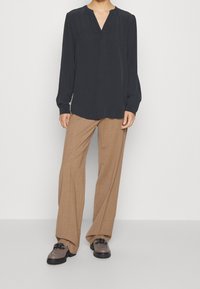 Sort bluse med V-hals og lange ærmer, parret med beige vidde bukser. Footwear har en mørk sål og dekorative snørebånd.