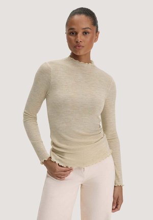 Frau trägt ein beiges, eng anliegendes Langarmshirt mit Rüschensaum und helle Hosen, steht vor schlichtem Hintergrund.