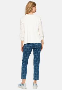 Cream long-sleeve top, relaxed fit; blue patterned jeans with subtle texture; white sneakers with blue accents; casual outfit from back view.