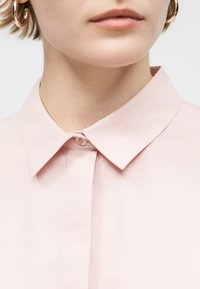 Κοντινό πλάνο ατόμου που φοράει ανοιχτό ροζ πουκάμισο με το πάνω κουμπί κλειστό και εμφανή χρυσούς κρίκους σκουλαρίκια.