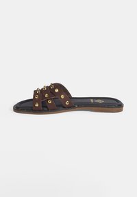 Bruine leren slipper met kruislings bevestigde bandjes en gouden studs, met een platte zool en een getextureerde zwarte binnenzool.