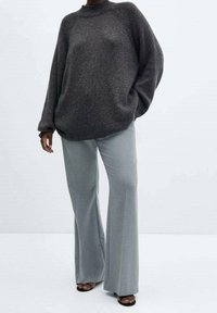 Persoon draagt een loszittende donkergrijze trui en lichtgrijze broek met wijde pijpen en open zwarte sandalen, staand tegen een effen witte achtergrond.