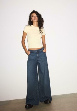Kvinde med krøllet hår står med hænderne i lommerne iført en lysegul kortærmet croptop og vide blå jeans med sorte sko.