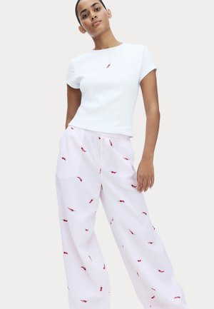 Mujer con camisa blanca de manga corta con un pequeño diseño de chile y pantalones blancos sueltos estampados con chiles rojos.
