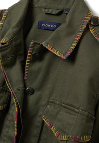 Giacca verde oliva con accenti colorati ricamati su colletto e tasche, bottoni neri e un'etichetta che riporta "PIOMBO". Realizzata in tessuto testurizzato.