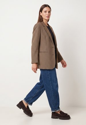 Frau geht und trägt einen braunen karierten Blazer, ein schwarzes Top, blaue hochgekrempelte Jeans und dunkelbraune Tassel-Loafers vor einfarbigem Hintergrund.
