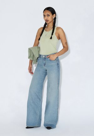 Hellgrünes Tanktop, hoch taillierte hellblaue Jeans mit weitem Bein und ein grüner Pullover liegt über dem Arm. Das Model hat schwarze Zöpfe und silbernen Schmuck.