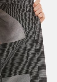 Tessuto a righe nere e grigie con una mano che tiene il bordo del capo, rivelando una texture liscia e un blocco di colore a contrasto nella parte inferiore.