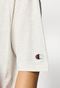 Ανοιχτό γκρι μπλουζάκι με κοντά μανίκια, με μαύρο και κόκκινο κεντημένο λογότυπο στο αριστερό μανίκι. Ομαλή υφή, casual εφαρμογή.