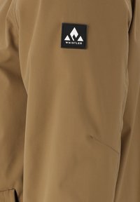 Bruine waterdichte jas met een gladde textuur, voorzien van een zwart vierkant label met de tekst "WHISTLER" en een geometrisch logo.