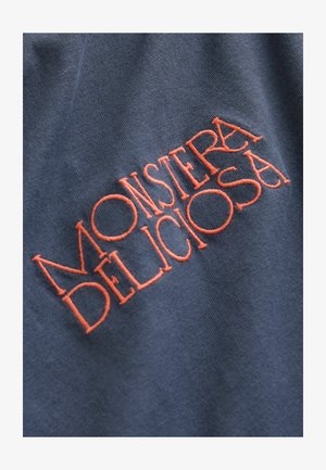 Marineblaues T-Shirt aus Stoff mit rotem, gesticktem Text, der "MONSTERA DELICIOSA" liest und eine glatte Textur sowie ein einfaches Design betont.