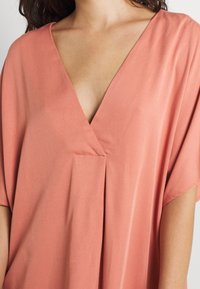 Pfirsichfarbene Bluse mit tiefem V-Ausschnitt, kurzen Ärmeln und lockerem, fließendem Design. Hergestellt aus weichem, leichtem Stoff mit glatter Textur.