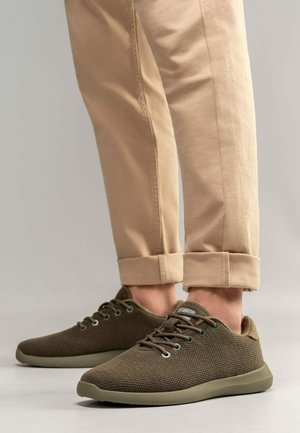 Baskets vert olive avec un dessus en maille texturée, lacets ronds et semelle en caoutchouc. Associées à un pantalon kaki retroussé.