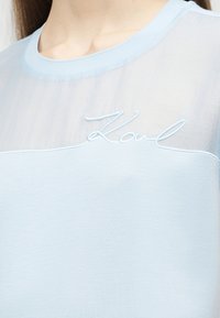 Světlomodrý průhledný top s kulatým výstřihem a nápisem "Karl" vyšitým kurzívou na horní části hrudi.